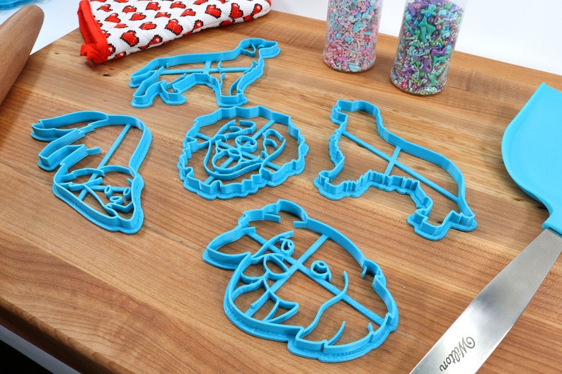 May include: A set of five blue plastic dog-shaped cookie cutters on a wooden surface. The cutters are in the shapes of a dog's head, a dog's body, and a dog's paw.