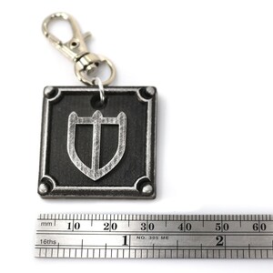 May include: A silver keychain with a black square charm featuring a silver shield design. The charm is approximately 3 cm wide.