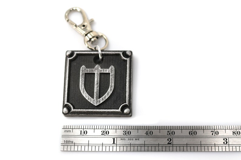 May include: A silver keychain with a black square charm featuring a silver shield design. The charm is approximately 3 cm wide.
