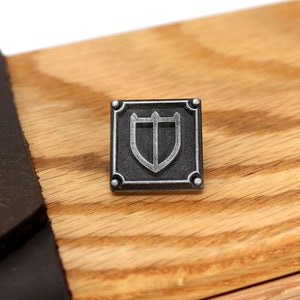 Puede incluir: Un emblema de escudo de metal plateado con fondo negro y forma cuadrada. El escudo tiene una parte superior puntiaguda y una parte inferior redondeada. El emblema est&aacute; sobre una superficie de madera.
