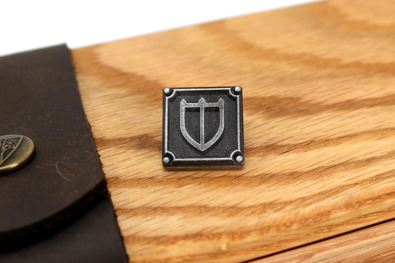 Puede incluir: Un emblema de escudo de metal plateado con fondo negro y forma cuadrada. El escudo tiene una parte superior puntiaguda y una parte inferior redondeada. El emblema est&aacute; sobre una superficie de madera.