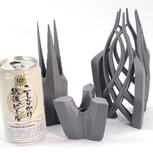 May include: Three 3D printed gray objects, one resembling a tall, pointed structure, another a curved, open cage, and the third a flat, abstract shape. A can of Japanese beer with the text "こしひかり" and "生ビール" is in the foreground.