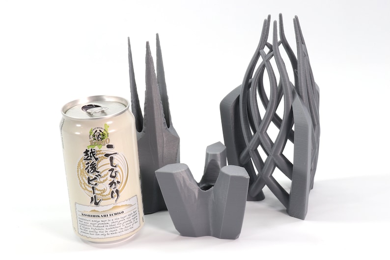May include: Three 3D printed gray objects, one resembling a tall, pointed structure, another a curved, open cage, and the third a flat, abstract shape. A can of Japanese beer with the text "こしひかり" and "生ビール" is in the foreground.