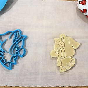 May include: Two cookie cutters, one blue and one beige, shaped like a cartoon character with a large hat and a bow. The cutters are on a white parchment paper.