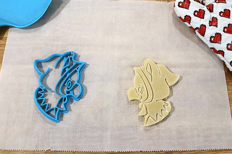May include: Two cookie cutters, one blue and one beige, shaped like a cartoon character with a large hat and a bow. The cutters are on a white parchment paper.