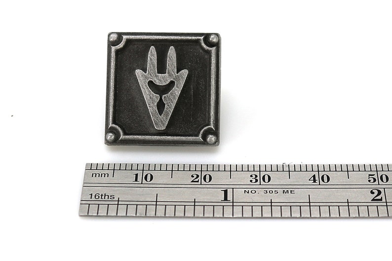 Op de afbeelding: Een zilverkleurige metalen pin met een gestileerd, drietandvormig symbool in een vierkant frame. De pin is ongeveer 2 cm breed.