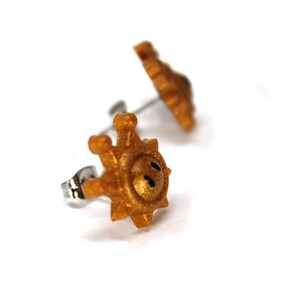 May include: A pair of gold-coloured earrings shaped like a sun with a smiley face. The earrings are made of metal and have a post back.