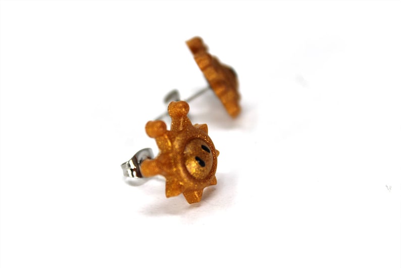 May include: A pair of gold-coloured earrings shaped like a sun with a smiley face. The earrings are made of metal and have a post back.