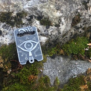 May include: A silver metal charm featuring a stylised eye design and a circuit board pattern. The charm is resting on a mossy rock.