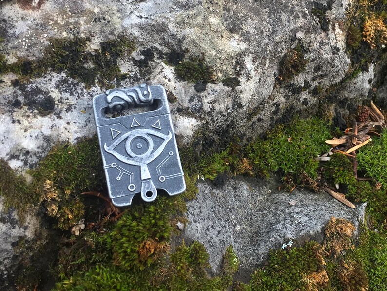 May include: A silver metal charm featuring a stylised eye design and a circuit board pattern. The charm is resting on a mossy rock.