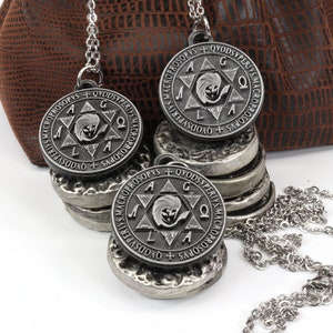 May include: Three silver-toned pendant necklaces with a circular design featuring text and a central emblem. The pendants rest on a stack of matching silver discs. A brown pouch is in the background.