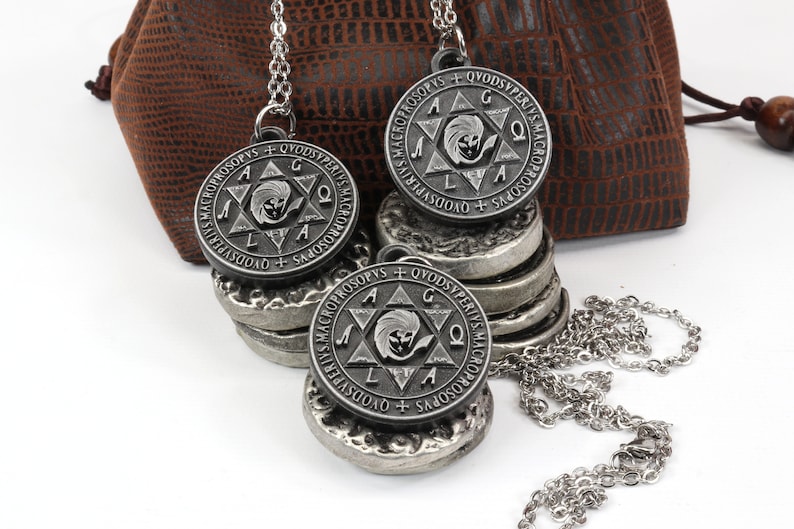 May include: Three silver-toned pendant necklaces with a circular design featuring text and a central emblem. The pendants rest on a stack of matching silver discs. A brown pouch is in the background.