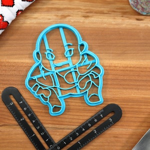 May include: A blue plastic cookie cutter shaped like a cartoon character. The character is wearing a shirt with a grid pattern and has a bald head.
