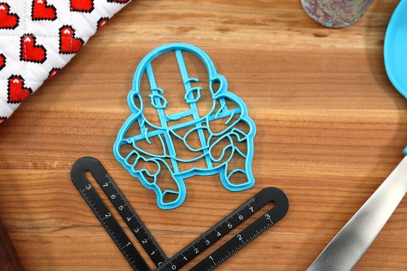 May include: A blue plastic cookie cutter shaped like a cartoon character. The character is wearing a shirt with a grid pattern and has a bald head.