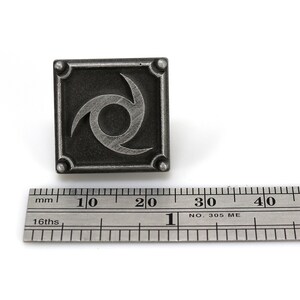 May include: A silver metal pin featuring a stylised black and white swirl design. The pin is approximately 2 cm wide and is shown next to a ruler.