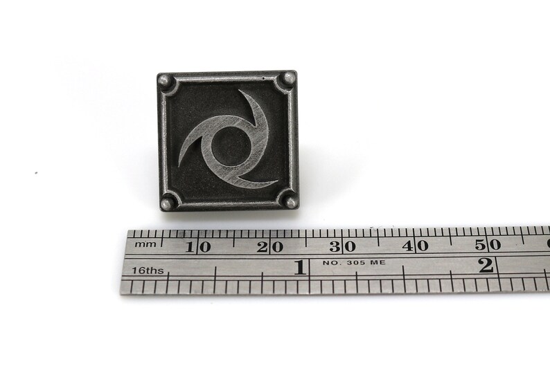 May include: A silver metal pin featuring a stylised black and white swirl design. The pin is approximately 2 cm wide and is shown next to a ruler.