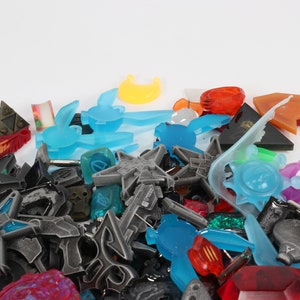 May include: A collection of colorful plastic charms in various shapes and sizes, including stars, moons, animals, and geometric designs. The charms are mostly blue, orange, and gray.