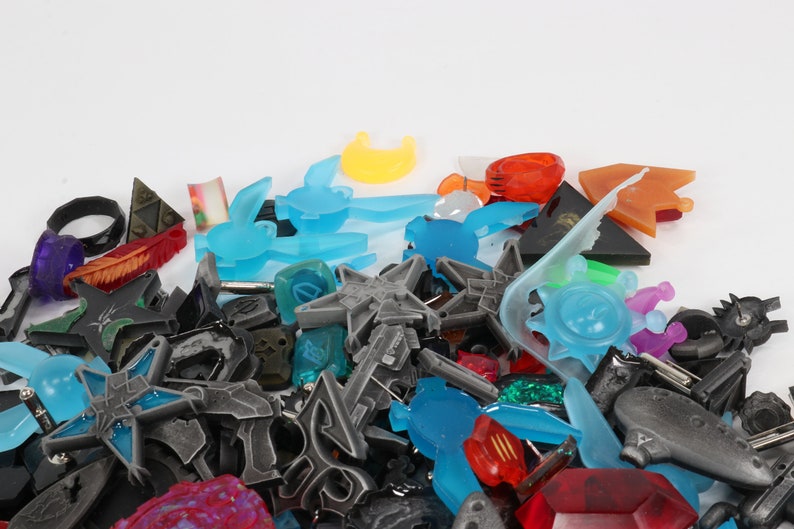 May include: A collection of colorful plastic charms in various shapes and sizes, including stars, moons, animals, and geometric designs. The charms are mostly blue, orange, and gray.