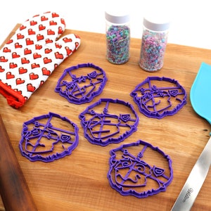 May include: Five purple plastic cookie cutters shaped like a dog's face, a rolling pin, a blue spatula, and two jars of sprinkles on a wooden surface.
