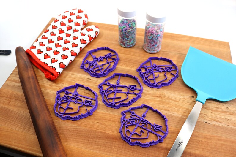 May include: Five purple plastic cookie cutters shaped like a dog's face, a rolling pin, a blue spatula, and two jars of sprinkles on a wooden surface.