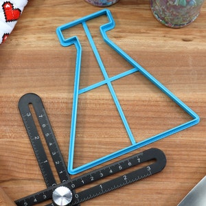 May include: A blue plastic cookie cutter shaped like a beaker with a grid pattern inside. The cutter is on a wooden surface with a ruler and other baking tools.