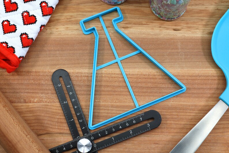 May include: A blue plastic cookie cutter shaped like a beaker with a grid pattern inside. The cutter is on a wooden surface with a ruler and other baking tools.