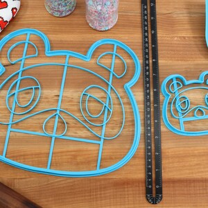 May include: Two blue plastic cookie cutters shaped like the face of a cartoon animal with large eyes and a nose. The larger cutter is about 6 inches wide and the smaller cutter is about 3 inches wide. The cutters are on a wooden surface.