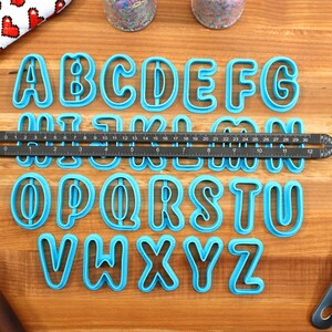 May include: A set of 26 blue plastic alphabet cookie cutters on a wooden surface. The cutters are arranged in alphabetical order. A ruler is placed below the first row of cutters.