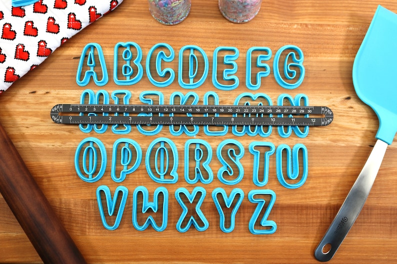 May include: A set of 26 blue plastic alphabet cookie cutters on a wooden surface. The cutters are arranged in alphabetical order. A ruler is placed below the first row of cutters.