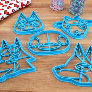 May include: Five blue plastic cookie cutters shaped like animal faces. The cutters are on a wooden surface.