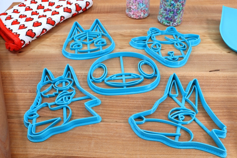 May include: Five blue plastic cookie cutters shaped like animal faces. The cutters are on a wooden surface.
