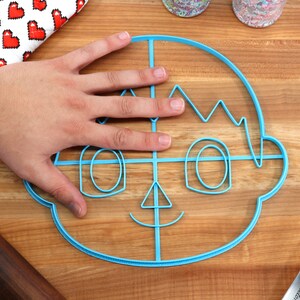 May include: A blue plastic cookie cutter shaped like a cartoon face with a smile, eyes, and hair. The cutter is on a wooden surface.