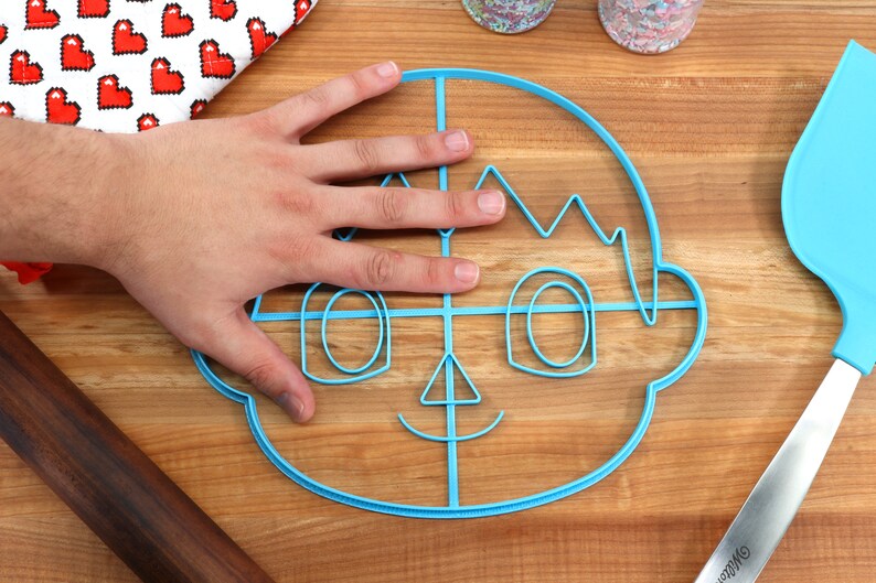 May include: A blue plastic cookie cutter shaped like a cartoon face with a smile, eyes, and hair. The cutter is on a wooden surface.