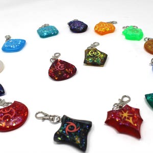 May include: A collection of 14 colorful resin keychains with different symbols and glitter accents. The keychains are arranged in a circle on a white background.