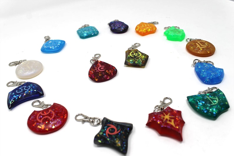 May include: A collection of 14 colorful resin keychains with different symbols and glitter accents. The keychains are arranged in a circle on a white background.
