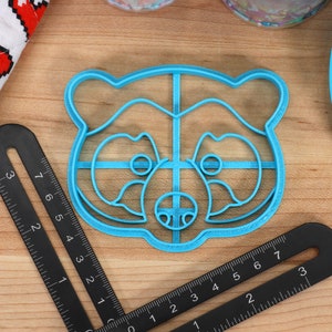 May include: A blue plastic cookie cutter shaped like a raccoon face. The cutter has a grid pattern inside the outline of the raccoon.