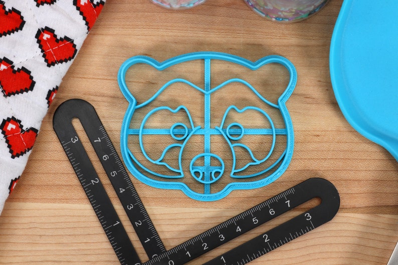 May include: A blue plastic cookie cutter shaped like a raccoon face. The cutter has a grid pattern inside the outline of the raccoon.