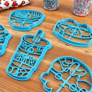 May include: A collection of blue plastic cookie cutters in various shapes, including a fish, a bubble tea cup with Chinese characters, a bowl of ramen, and a heart. The cutters are arranged on a wooden surface, with sprinkles in the background.