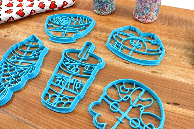 May include: A collection of blue plastic cookie cutters in various shapes, including a fish, a bubble tea cup with Chinese characters, a bowl of ramen, and a heart. The cutters are arranged on a wooden surface, with sprinkles in the background.