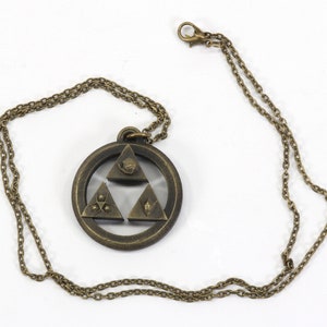 Puede incluir: Un collar dorado con un colgante que presenta el s&iacute;mbolo de la Trifuerza de la serie de videojuegos The Legend of Zelda. El colgante es de dise&ntilde;o circular con el s&iacute;mbolo de la Trifuerza en el centro, y la cadena es de estilo de eslab&oacute;n simple.