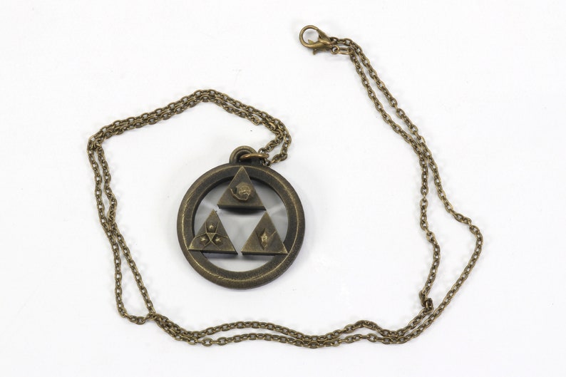 Puede incluir: Un collar dorado con un colgante que presenta el s&iacute;mbolo de la Trifuerza de la serie de videojuegos The Legend of Zelda. El colgante es de dise&ntilde;o circular con el s&iacute;mbolo de la Trifuerza en el centro, y la cadena es de estilo de eslab&oacute;n simple.
