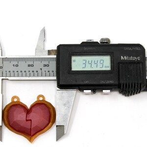 以下が含まれることがあります： 赤と金のハート型のペンダントをデジタルノギスで測定しています。ノギスは34.49mmを示しています。