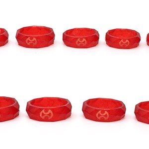 Op de afbeelding: Tien gefacetteerde, rode harsringen met een goudkleurig embleem. De ringen zijn gerangschikt in twee rijen van vijf tegen een witte achtergrond. De ringen hebben een geometrisch ontwerp.