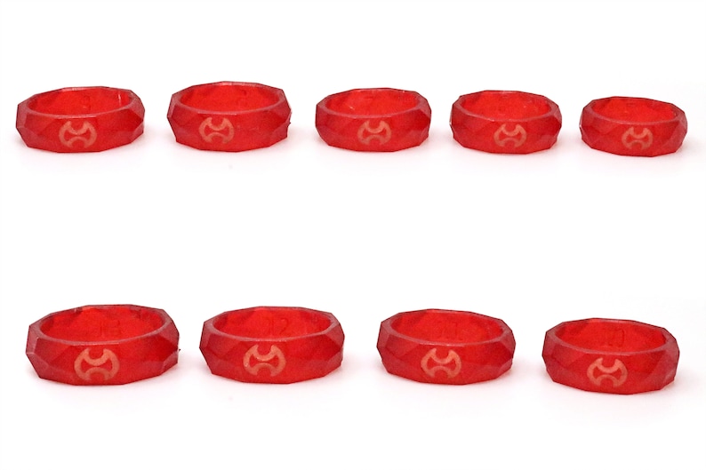 Op de afbeelding: Tien gefacetteerde, rode harsringen met een goudkleurig embleem. De ringen zijn gerangschikt in twee rijen van vijf tegen een witte achtergrond. De ringen hebben een geometrisch ontwerp.