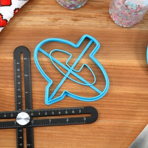May include: A light blue cookie cutter in the shape of a speech bubble with a line through it, a black and silver measuring tool, and a blue spatula on a wooden surface. A white cloth with red pixelated hearts is in the background.