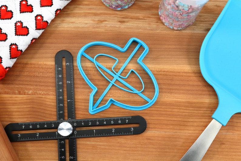 May include: A light blue cookie cutter in the shape of a speech bubble with a line through it, a black and silver measuring tool, and a blue spatula on a wooden surface. A white cloth with red pixelated hearts is in the background.