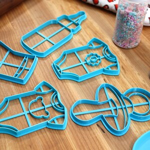 May include: Five blue plastic cookie cutters shaped like a baby bottle, a baby onesie with a flower, a baby onesie with a bear, a baby onesie with a flower, and a pacifier. The cutters are on a wooden surface.
