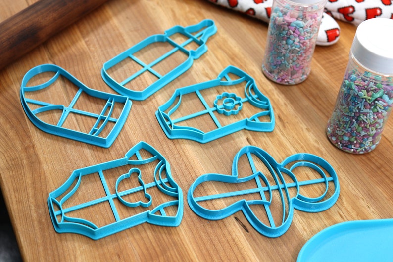 May include: Five blue plastic cookie cutters shaped like a baby bottle, a baby onesie with a flower, a baby onesie with a bear, a baby onesie with a flower, and a pacifier. The cutters are on a wooden surface.