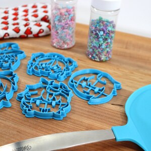 K&ouml;nnte beinhalten: Blaue Plastik-Ausstecher in Form eines Weihnachtsbaums, eines Schneemanns und eines Rentiers. Die Ausstecher liegen auf einer Holzoberfl&auml;che mit zwei Gl&auml;sern Streusel im Hintergrund.