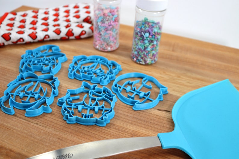 K&ouml;nnte beinhalten: Blaue Plastik-Ausstecher in Form eines Weihnachtsbaums, eines Schneemanns und eines Rentiers. Die Ausstecher liegen auf einer Holzoberfl&auml;che mit zwei Gl&auml;sern Streusel im Hintergrund.